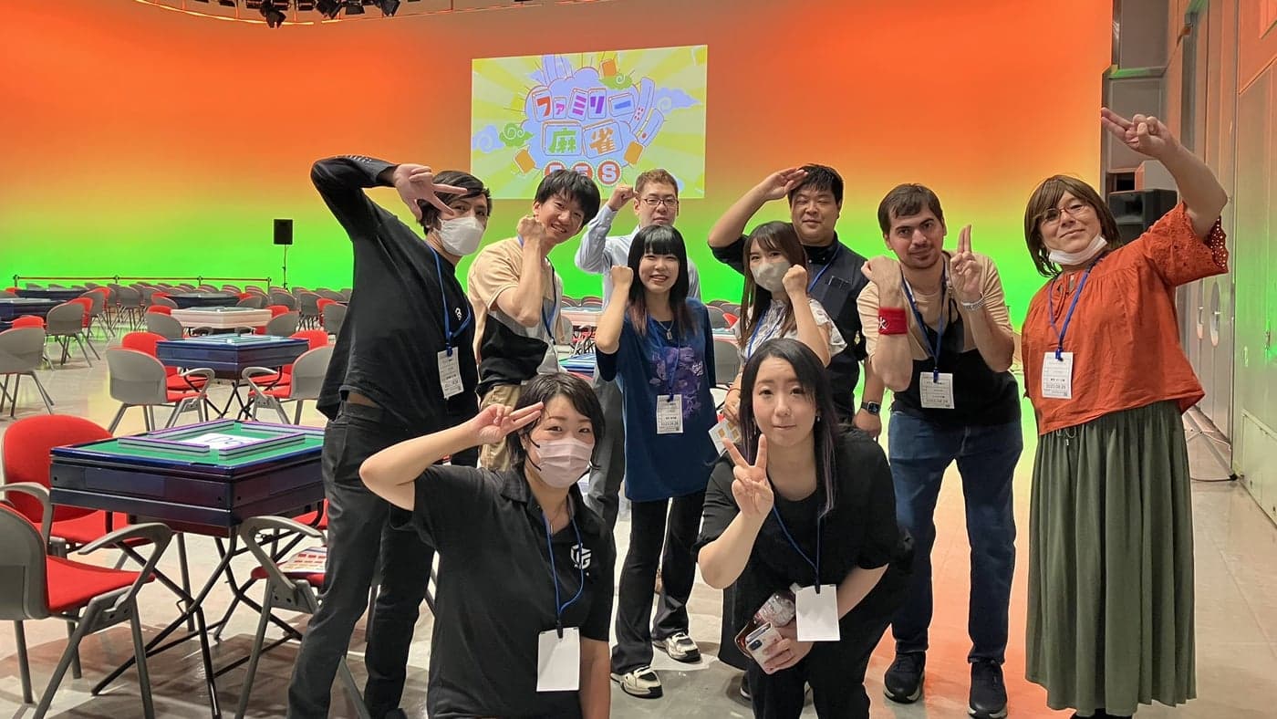 「ファミリー麻雀FES」の映るスクリーンの前でポーズをとる運営スタッフの集合写真。後列の右から2人目、赤いリストバンドの男性がニコラス