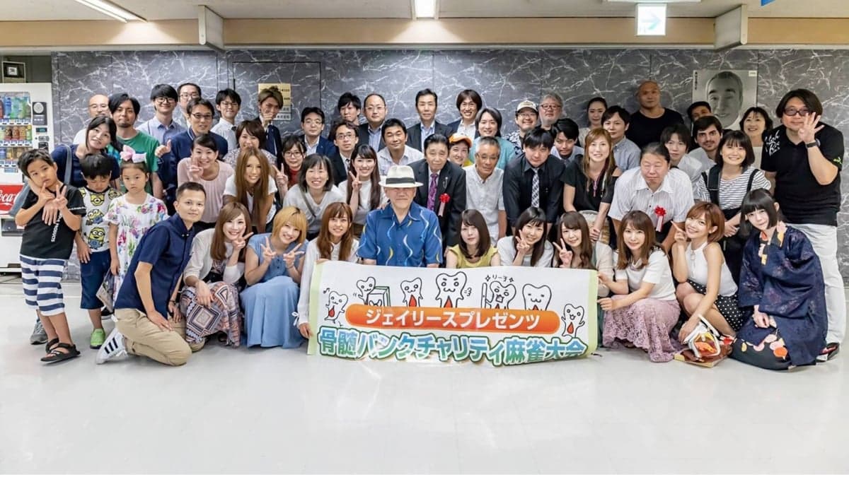 「ジェイリースプレゼンツ 骨髄バンクチャリティ麻雀大会」の横断幕を前に並ぶ、大会に集まった大勢の人たちの集合写真